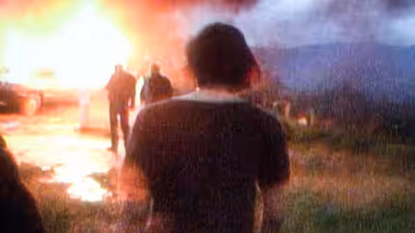 A young man watches a burning vehicle with others in a rural area