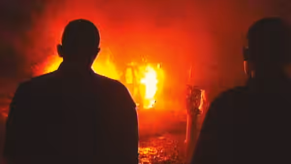 Two men stand silhouetted against a burning car in the darkness