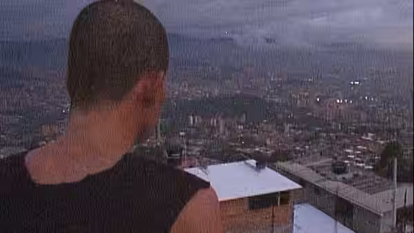 A man with short hair gazes over a city from a rooftop at dusk