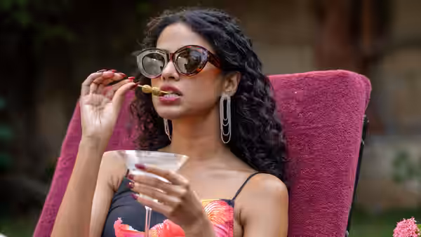 A woman wearing sunglasses eats from a skewer while seated outdoors with a drink