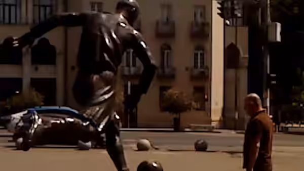 A bald man standing beside a large bronze soccer statue in a square