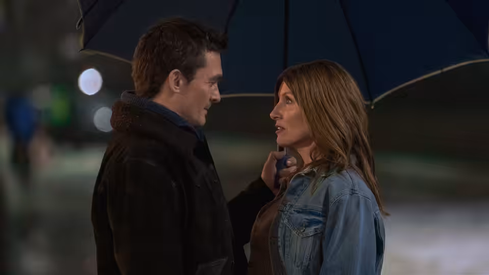 Sharon Horgan and Rupert Friend stand close under a blue umbrella at night