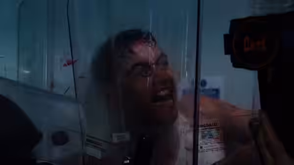 A shirtless man grimaces with his face pressed against a glass partition