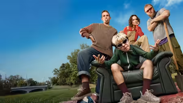 A boy in sunglasses gestures from a green armchair as three adults surround him outdoors