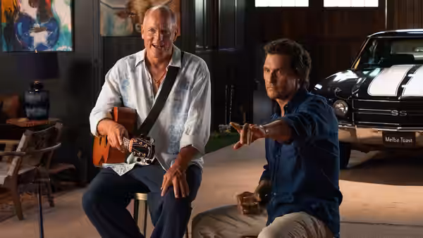Woody Harrelson plays guitar indoors while Matthew McConaughey gestures beside him
