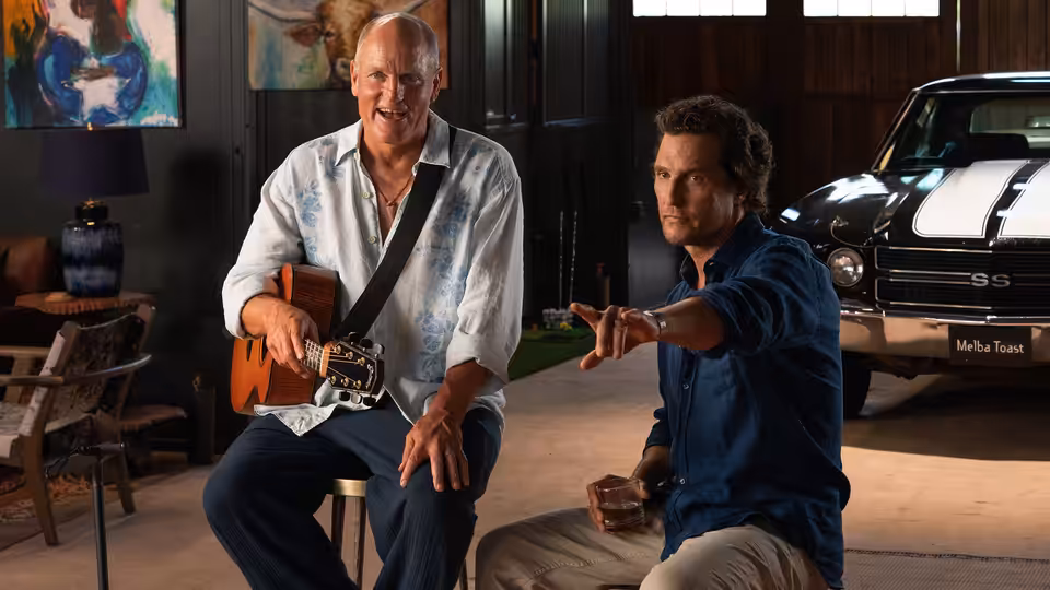 Woody Harrelson plays guitar indoors while Matthew McConaughey gestures beside him
