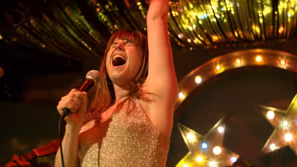 Rose-Lynn (Jessie Buckley) sings into a microphone beneath stage lights, one arm raised