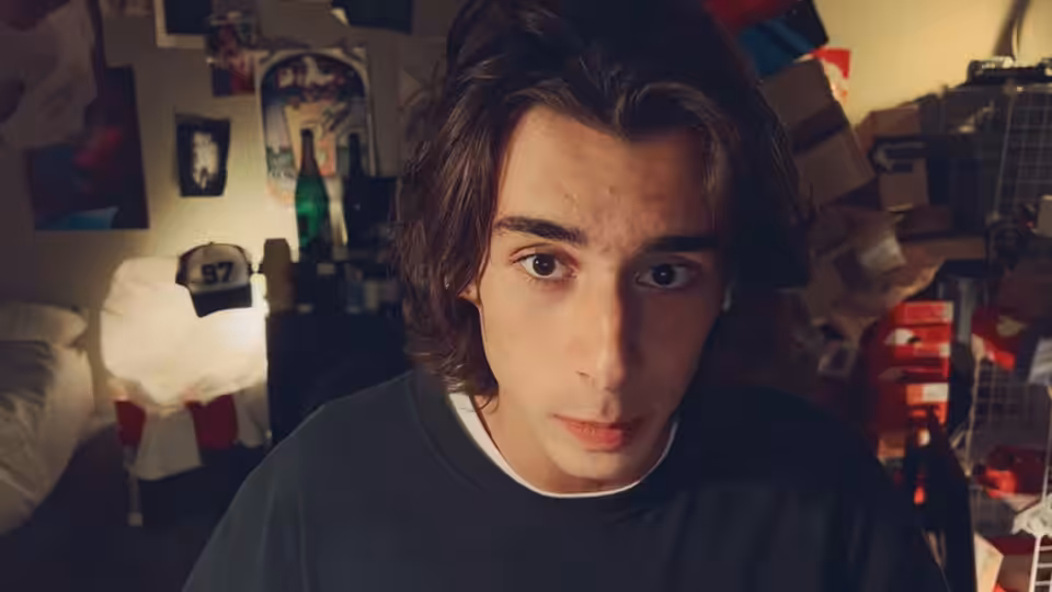 a young man with brown hair stares ahead in a cluttered bedroom with posters and boxes
