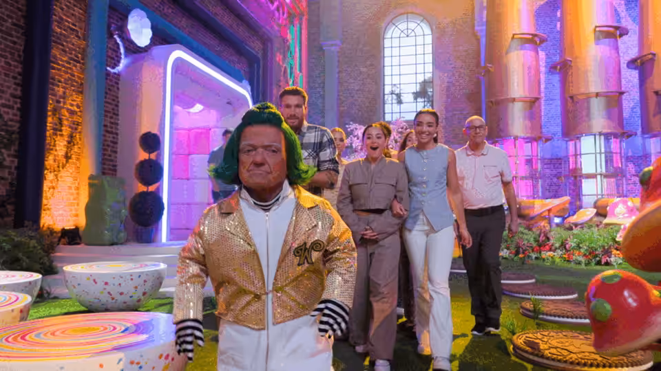 A man in a gold jacket stands before contestants in a brightly colored game-show room
