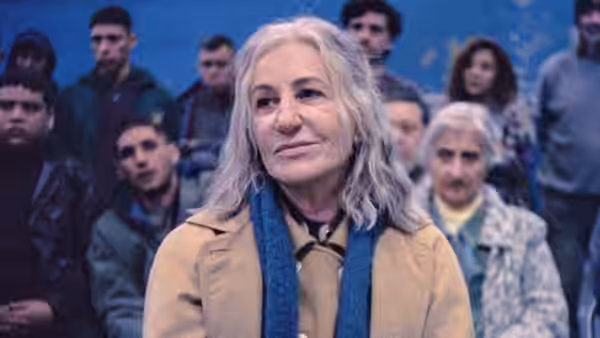 A silver-haired woman in a tan coat and blue scarf stands among a crowd