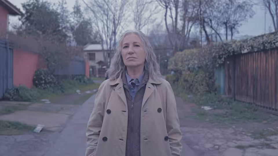 A gray-haired woman in a beige coat stands on a paved path among bare trees