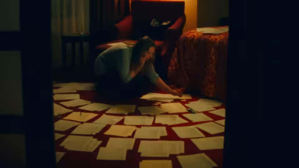 Man kneels on red rug amid scattered papers holding his head