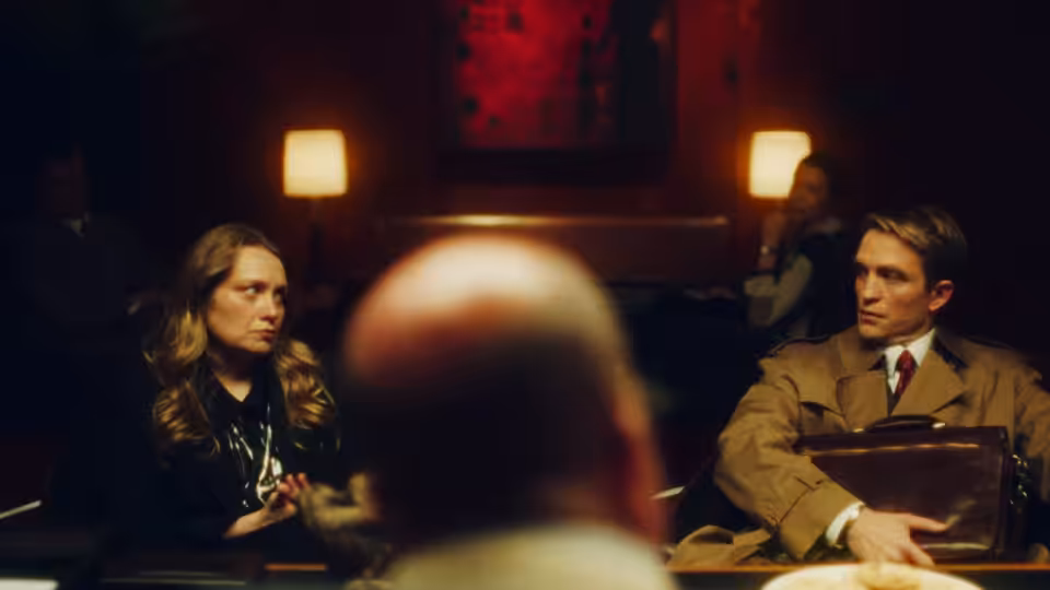 Man in trench coat seated beside woman at table in dark room
