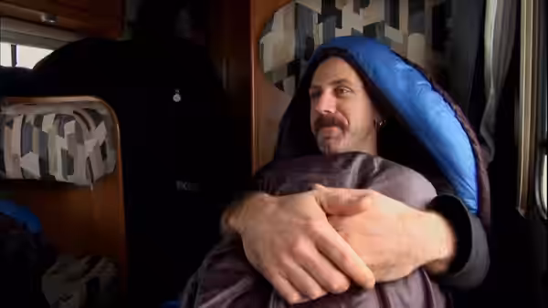 Man with mustache wrapped in blue sleeping bag sitting inside a camper van
