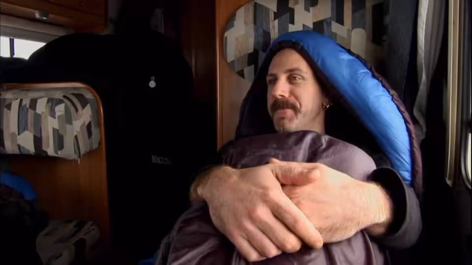 Man with mustache wrapped in blue sleeping bag sitting inside a camper van