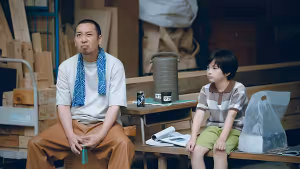 Man and boy sit side by side on a bench inside a cluttered workshop