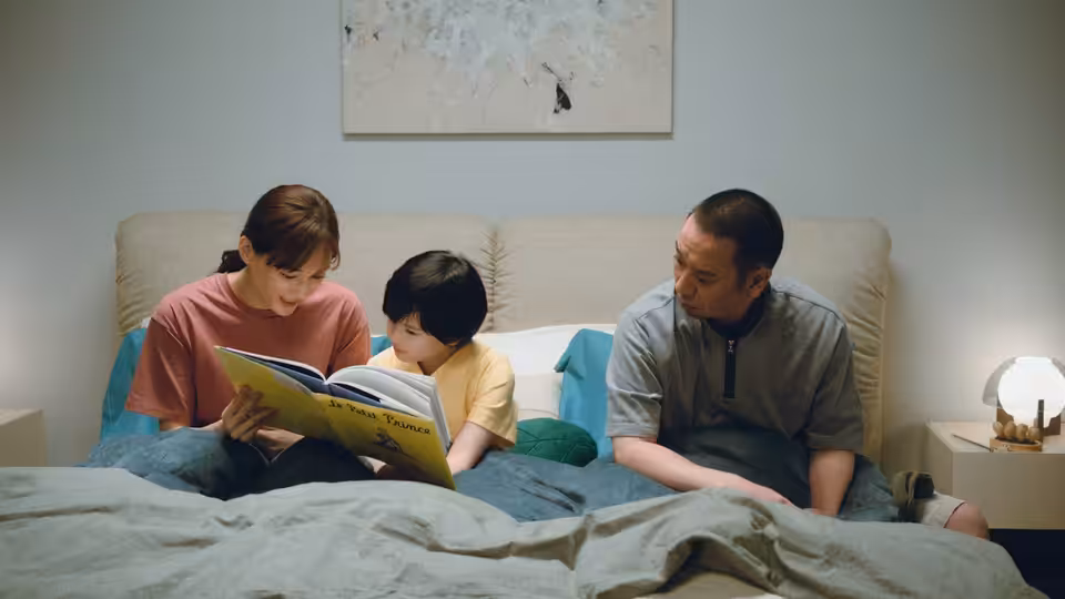 Woman reads a book aloud to a boy as a man watches from the bed