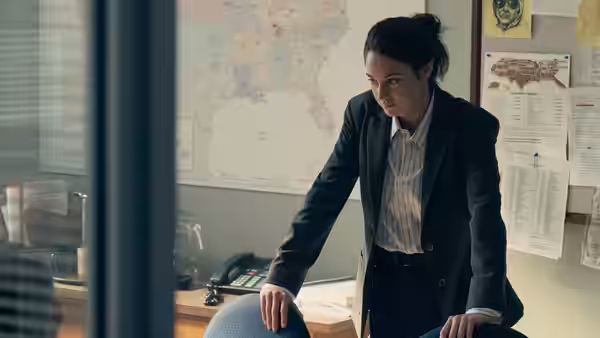 Woman in dark suit leans on desk in office with maps on the wall