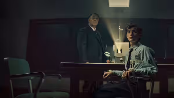 A young man sits at a desk as an older man stands behind it in a dim office