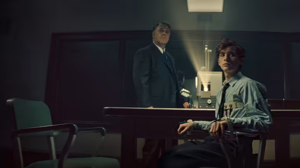 A young man sits at a desk as an older man stands behind it in a dim office