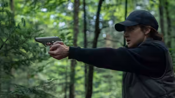 Woman in cap and vest aims a handgun through dense green forest