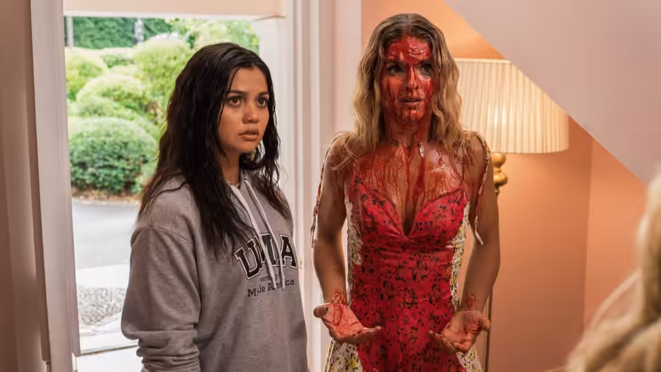 Nora (Siena Agudong) stands beside a blood-covered woman in a home's entryway