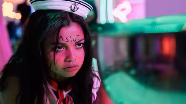 A dark-haired woman in a sailor hat, with an insult written on her forehead, looks ahead