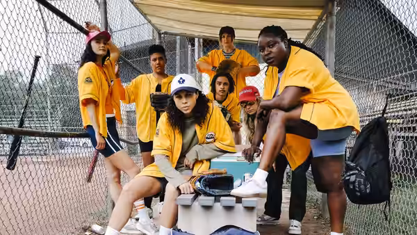 A softball team in yellow jerseys gathers around a cooler in a fenced dugout
