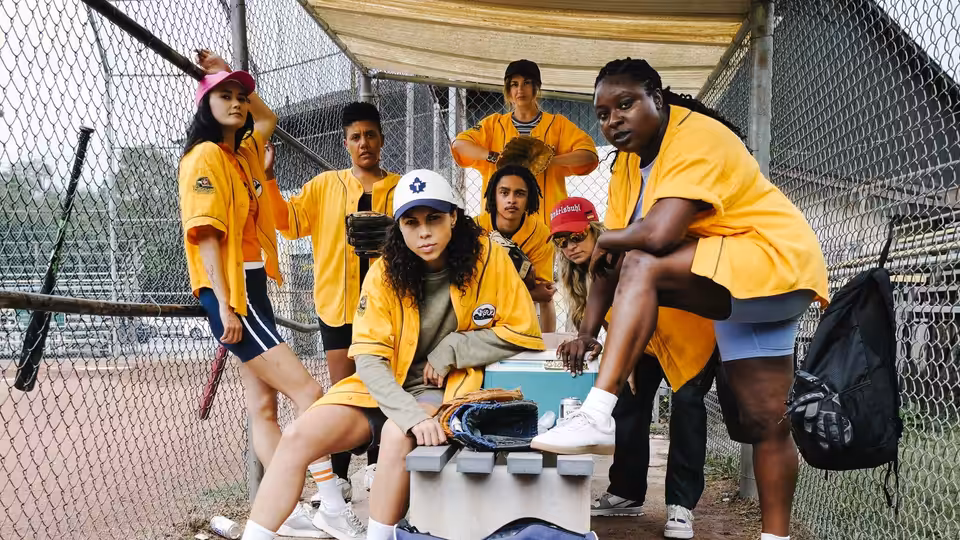 A softball team in yellow jerseys gathers around a cooler in a fenced dugout