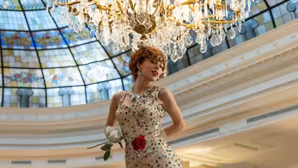 A woman in an embroidered gown holds a red rose beneath a chandelier