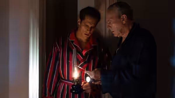 Two men in robes inspecting an object with lantern and flashlight in a dark doorway