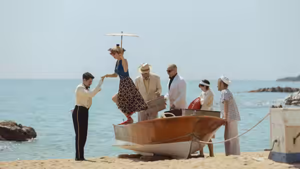 Woman in polka-dot dress stepping into a wooden boat on the beach with help