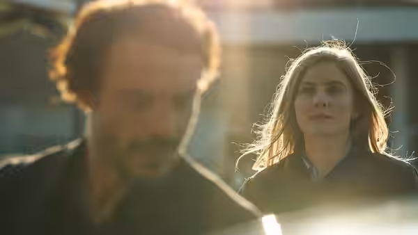 A woman watches a man walking ahead of her in a sunlit courtyard