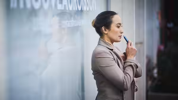 A woman in a taupe coat checks her reflection in glass while applying lipstick