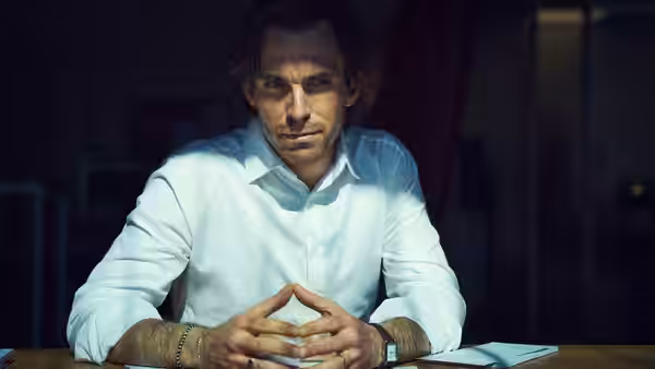 A man sits at a table with his hands clasped in a dim room