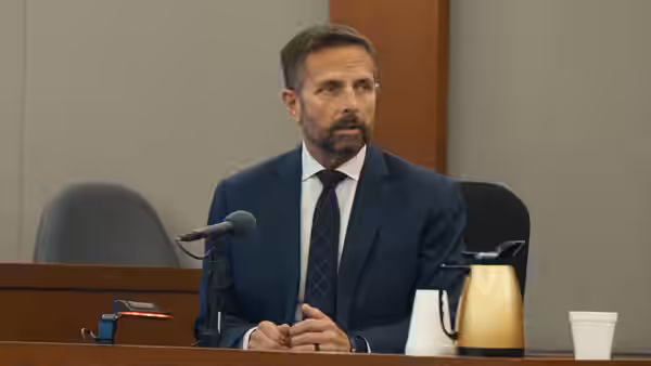 A bearded man in a suit sits at a courtroom table beside a microphone