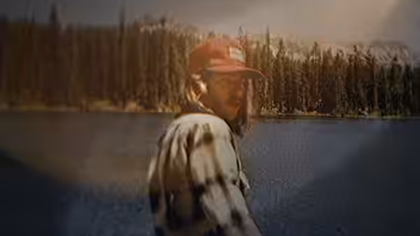 A man in a cap turns toward the camera beside a lake and forest