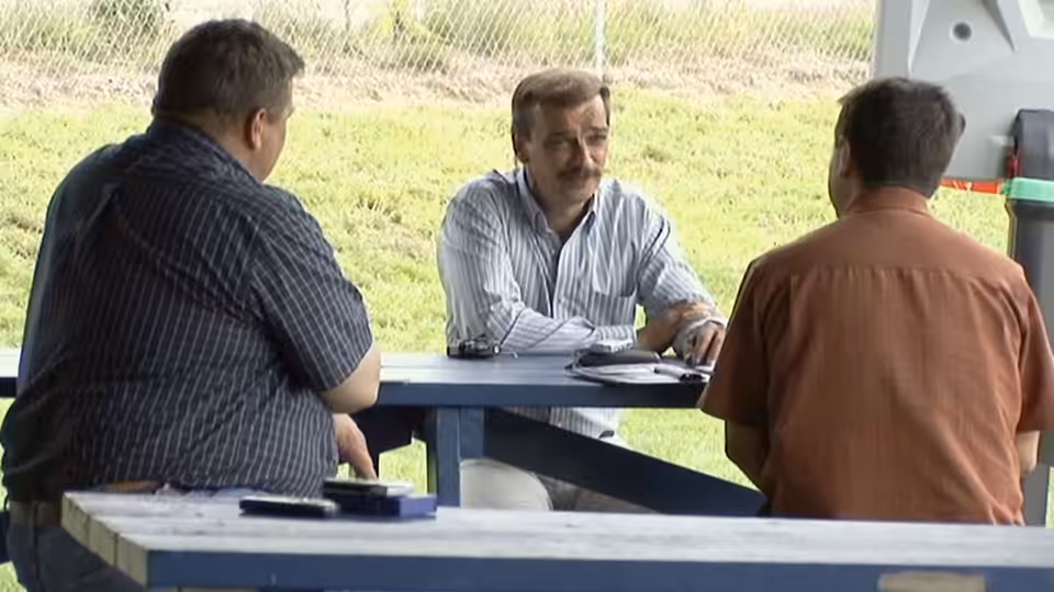 Three men sit around a picnic table outdoors, talking over papers and a plate
