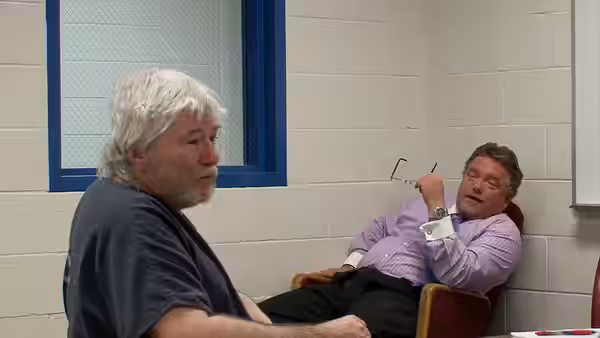 Two older men sit in a small room, one upright and the other reclining
