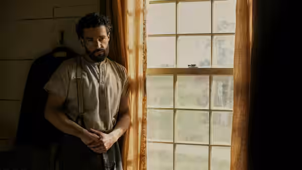A bearded man in suspenders standing by a window with hands clasped