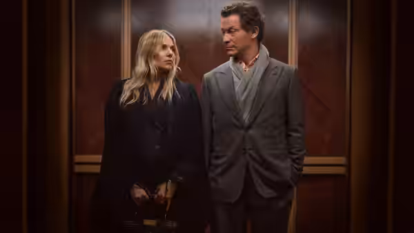 A blonde woman and a man in a suit stand in a wood-paneled room