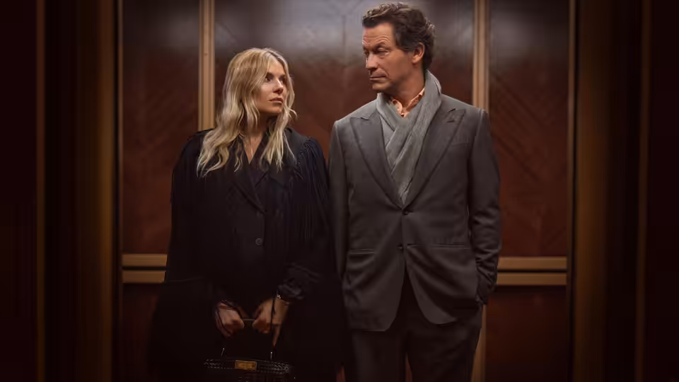 A blonde woman and a man in a suit stand in a wood-paneled room