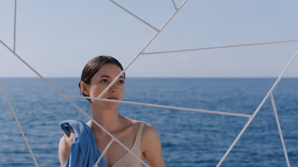 Cécile (Lily McInerny) holding blue cloth by the sea behind white structure