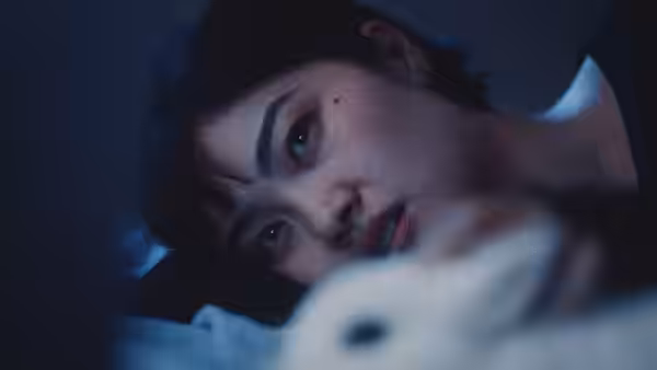 A young woman with short dark hair lies in bed beside a white stuffed rabbit