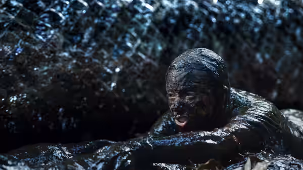 A mud-covered person struggles through dark water beneath a looming scaled creature