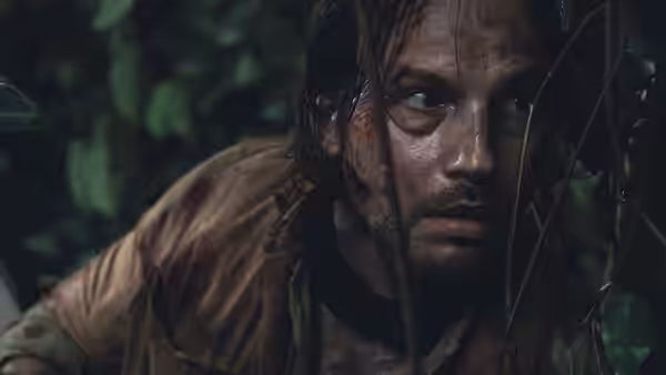 A bloodied man crouches among dense jungle foliage, watching something through the leaves