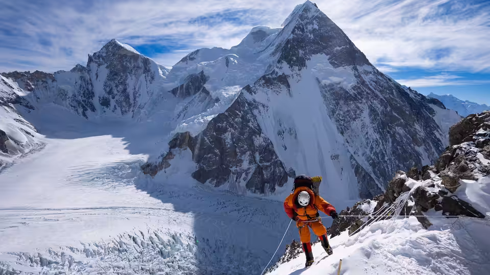 An expedition member in orange stands on a ridge above snowy peaks