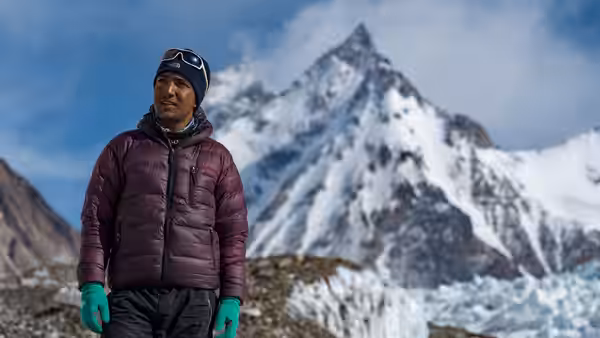 A mountaineer in winter gear stands before a snow-covered Himalayan peak