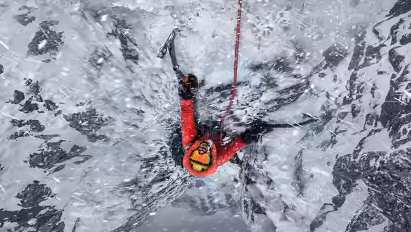 A climber in red suit ascends a sheer icy wall with axe and rope