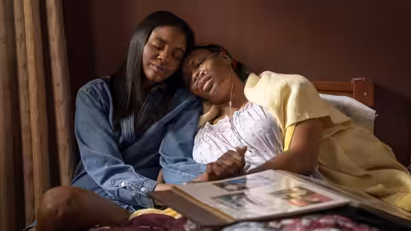 Two women sit together on a bed, one resting her head on the other's shoulder beside an open album